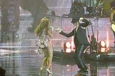 A woman and a man are dancing together on stage. The brunette female wears a short pink dress, and she sings. On the other side, he wears a dark suit.