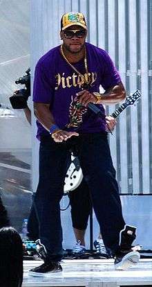 Flo Rida rapping in a concert, wearing a purple shirt, sweatpants, and a cap.