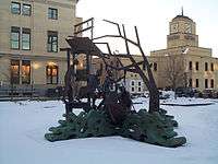 A metal statue showing several people getting into a boat. In the background are several buildings. Snow is seen on the ground.