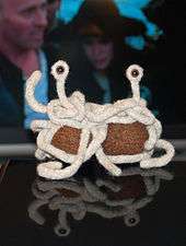 small handmade knit Flying Spaghetti Monster sitting on a table with people dressed as pirates in background.