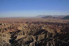 The shot is of a barren desert environment. There are several canyons, and it is framed against a blue sky.