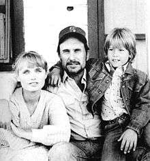 A black-and-white image of a bearded man wearing a baseball cap, sitting on the front porch of a wooden house, with his right arm around a smiling woman wearing a sweater, and his left arm around a smiling young boy sitting on his lap.