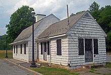 A small white building sided in shingles with a black asphalt bell-shaped roof, seen from its left. A slightly lower eing with a pointed roof projects toward the right. Its doors are screened and locked, and it looks neglected. Two black iron lamppots rise next to a curb and pavement at its left
