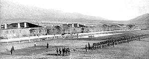 A black and white picture of Fort Grant near Stafford, Arizona circa 1885. Center middle ground to right middle ground is about four or five companies of soldiers standing in ranks. The buildings of the Fort in the background.