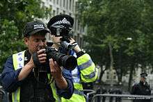 Two uniformed FIT police officers standing on the street, one has a camera with a large lens and then other has a small camcorder