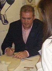 A dark-haired, middle-aged man in a black suit sits at a table, signing autographs.