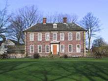 A two-storey brick-built mansion in its own well-kept garden of green grass. The ground floor has six windows – three on each side of a wooden front door. The second floor has seven windows. The house has a grey slate roof surmounted by two small brick chimneys. Behind the house are leafless trees and behind those, a clear blue sky.