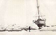 Front view of ship showing it listing to the right. It is surrounded by ice, but in front of the ship a narrow channel of open water is visible. The scene is watched by a man standing on the ice.