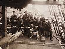A group of 13 men and one dog pose on the cramped deck of a ship, amid ropes, spars and rigging, all wearing hats and, with one exception, dark suits.