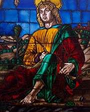 Detail of a stained glass window depicting a saint dressed in yellow, green and red robes, holding a quill.