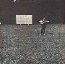 The album cover is a black-and-white photograph of a sports field with a score board in the distance, off-center to the left. Off-center to the right and also in the distance Fred Frith is standing playing a guitar. There is no text on the cover.