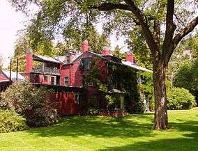 A large, rambling red house with dark trim stands in a green yard with a tree shading it.