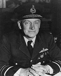 Half-portrait of uniformed man in peaked cap, with pilot's wings on left-breast pocket, clasping hands together