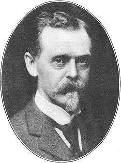 Black-and-white photograph, set in an oval frame, of the head and shoulders of a man of about 55. Dressed formally in a dark suit, dark tie, and white collar, he is looking directly out of the frame. He has dark eyes and dark hair, but his neatly trimmed moustache and goatee are gray.