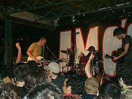 A color photograph of members of the band Fugazi performing on state with the heads of an audience in the foreground