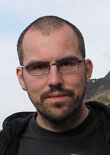 Head shot of a young man with glasses, dark brown buzz cut and stubble.