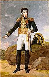 Formal full-length portrait of a Tharreau in uniform, in a rocky landscape. He is a short, solidly built man with a round face and alert dark eyes. His pale skin contrasts with his black curly hair and heavy cheek whiskers. He looks out of the picture to the left while gesturing backward with his right hand. He holds a sword in his left hand.