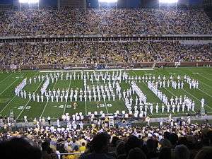 There is a large football stadium with yellow bleachers and green grass, seen at night. The band is on the field facing the viewer, wearing white uniforms, holding instruments, and arranged to form an interlocking GT. Many people are in the stands.
