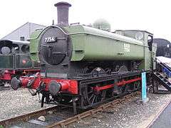 A pannier tank locomotive is on static display with wooden steps leading up to the cab. The cab, bunker, and pannier tank are painted light green. The chimney, smokebox, splashers, running plate, and wheels are all in black. The front buffer beam and coupling rods are red. The words "NCB 7754" are shown in white on the side of the pannier tank. The number is also shown on the smokebox door in white letters.
