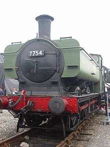 A pannier tank locomotive is seen from the front a slightly to the right. The cab and pannier tank are painted green. The chimney, smokebox, running plate, splashers, and wheels are black, and the buffer beam and coupling rods are red. The number, 7754, is shown in white letters on a black plate on the smokebox door. Part of round cab window is visible and is edged in brass. The white lettering "NCB 7754" is on the side of the pannier tank.