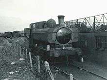 A pannier tank locomotive stands alone in a scrapyard next to a semi-derelict building. In the background are other locomotives waiting to be scrapped. To the left is a fence of concrete posts with wire.