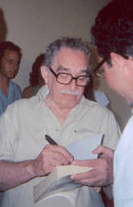 Grey haired man with a moustache and glasses stands while  autographing books.