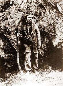 A man with beard and long hair is holding a long gun and is standing in front of a very large tree.