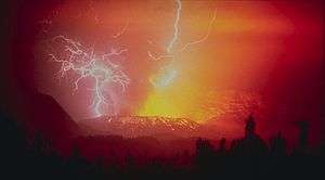 A photograph depicting lightning striking a volcano that is in the process of erupting bright yellow lava into the air, all surrounded by a red haze.