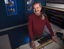 An older man with salt-and-pepper hair and a mustache stands smiling in a radio station. He is wearing a burgundy sweater.