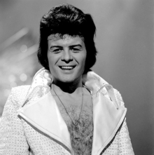 Black-haired man with a hairy chest, wearing a shiny jacket open to the waist, with large lapels, smiles towards the camera.