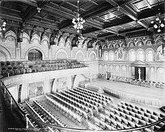 An auditorium with a balcony and raised stage surrounded by decorative arches on the walls.