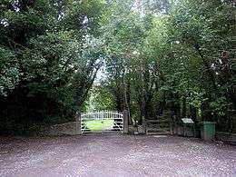 A white-painted metal double gate and a wooden pedestrian gate in a low stone wall, with a gravel carpark to the fore and a grassy area and trees to the rear.