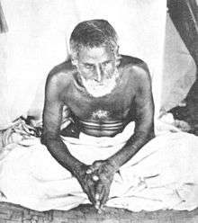 A photograph of a bearded Indian ascetic dressed in dhoti and sitting down cross-legged