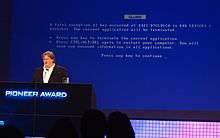Gabe Newell stands at a podium in front of a large screen. The screen is blue, similar to the "blue screen of death" for Windows, but with different text, including the word "GLaDOS".