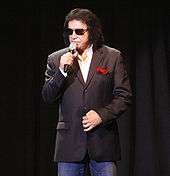 A man wearing a black pin-striped sports coat with a red Handkerchief, a white dress shirt, blue jeans, and sunglasses, and holding a microphone
