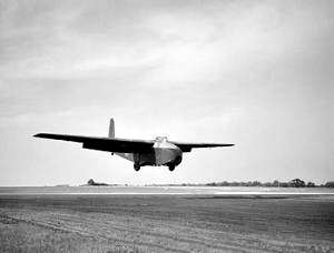 A large, bulky glider with long wings in flight, coming in to land on a runway. It is casting a shadow to its right on the runway. Fields can be seen behind it, and there are trees and bushes in the background.