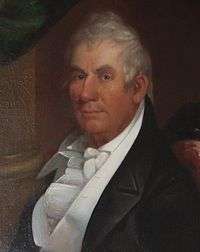 A man with gray hair and a ruddy complexion wearing a black jacket and frilly white vest