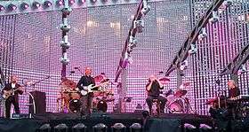 Four men on a stage; two are playing guitars, one is sitting on a stool and holding a microphone, and one is playing keyboards. Various stage equipment, lighting fixtures, drum sets, speakers and other audio equipment can be seen in the background.