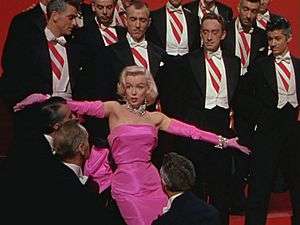 Monroe in Gentlemen Prefer Blondes. She is wearing a shocking pink dress with matching gloves and diamond jewellery, and is surrounded by men in tuxedos.