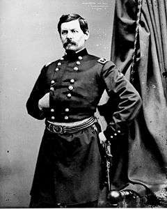 Man with moustache in uniform with two vertical columns of buttons with left hand on hip and right hand tucked inside uniform