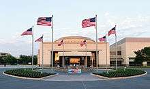 Above the doorway of a large, relatively plain rectangular structure with a short dome are the words "George Bush Library". In front of the building is a circular courtyard with a water fountain; eight American flags are positioned evenly around the circle.