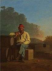 A man with graying hair and beard sits on a crate and leans against a barrel. He is wearing a red hat, white shirt, blue pants, and brown boots, and is smoking a pipe
