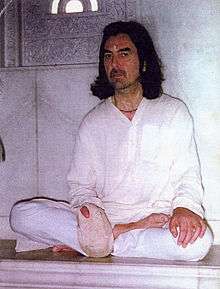 A photo of George Harrison sitting cross-legged with chanting beads in a bag on his right hand