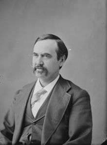 A man with dark hair and a mustache wearing a dark jacket and vest, light tie, and white shirt