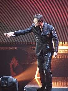 A man dressed in a black leather suit standing on a stage holding out a microphone to an audience