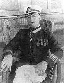 A white man sitting relaxed in a wicker chair. He is wearing a white cap, light–colored pants, and a dark jacket with four round medals on the left breast and a star–shaped medal around the neck.