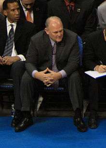 A man, wearing a black suit and a blue tie, is watching the basketball game while sitting courtside.