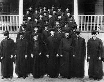 Thirty-four men in black robes and square academic caps stand on an outdoor staircase.