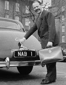 Full length picture of a middle aged man with a very large, bushy mustache. He is wearing a suit and carrying a briefcase. He is standing at the rear of a parked car with his hand on the boot-handle.