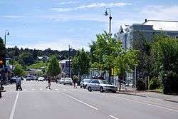 A busy small-town straight street, flanked by shops and the occasional small tree. In the foreground is a pedestrian crossing. Three people are crossing right-to-left - two ladies stepping off the crossing and another entering it. Another lady is crossing left-to-right about halfway across. In the background is a Shell petrol station and behind that a small hill with a large house about halfway up.